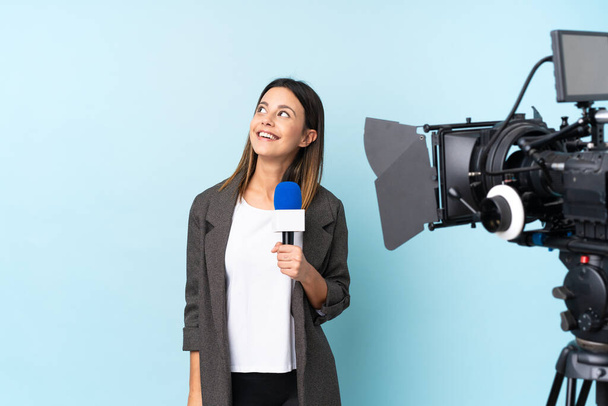 Женщина-репортер держит микрофон и сообщает новости на изолированном синем фоне смеясь и глядя вверх
 - Фото, изображение