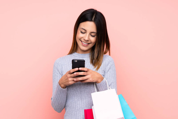 Woman over isolated pink background holding shopping bags and writing a message with her cell phone to a friend - Photo, image