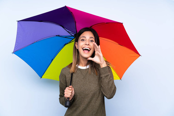 Молода брюнетка жінка тримає парасольку над ізольованою синьою стіною кричить з відкритим ротом
 - Фото, зображення