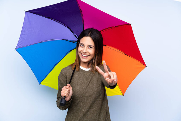 Молодая брюнетка с зонтиком над изолированной голубой стеной улыбается и показывает знак победы
 - Фото, изображение