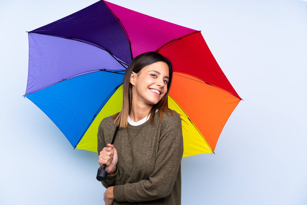 jonge brunette vrouw holding een paraplu over geïsoleerde blauwe muur lachen - Foto, afbeelding