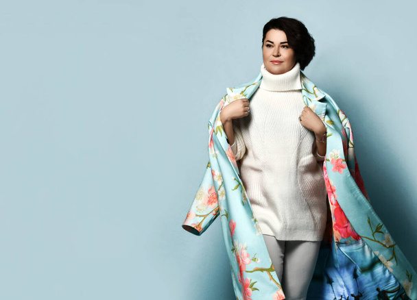 Brunette girl in white sweater and jeans, coat with floral print. Smiling, holding coat with her hands, posing on blue background - Photo, Image
