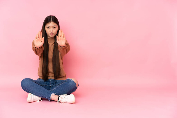 Fiatal ázsiai nő ül a padlón elszigetelt rózsaszín háttér így stop gesztus és csalódott - Fotó, kép