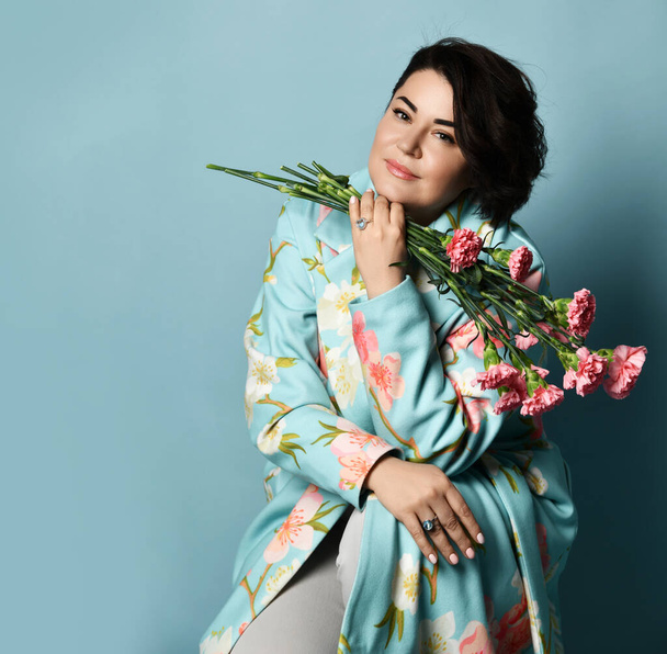 Female in white jeans and coat with floral print. Holding bouquet of pink carnations, sitting against blue studio background - Foto, imagen