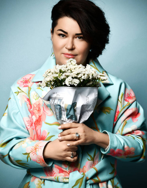 Female in coat with floral print. Smiling, holding bouquet wrapped in paper with project of apartment, posing on blue background - Foto, Imagen