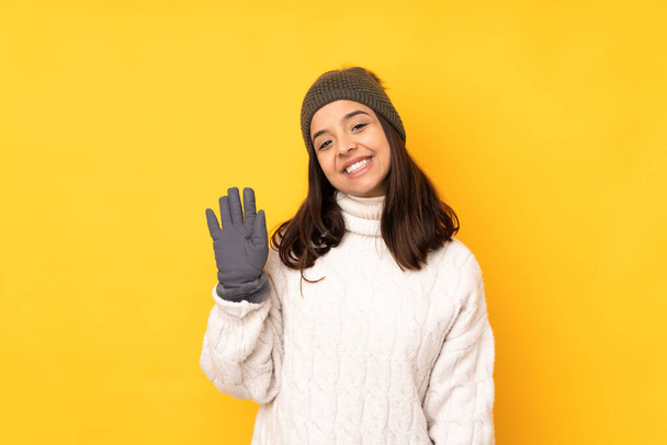 Mujer joven con sombrero de invierno sobre fondo amarillo aislado saludando con la mano con expresión feliz
 - Foto, Imagen