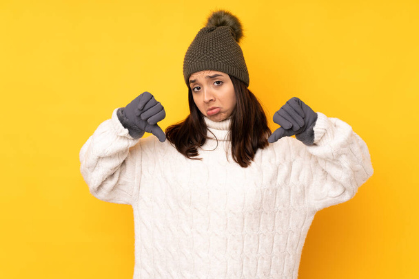 Молода жінка з зимовим капелюхом над ізольованим жовтим тлом показує великий палець вниз
 - Фото, зображення