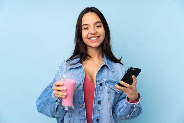 Young woman with strawberry milkshake over isolated blue background holding coffee to take away and a mobile - Photo, Image