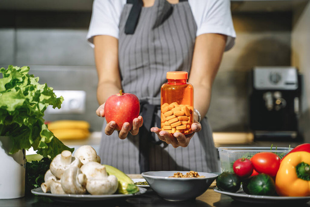 Close up of female hands holding apple and nutritional supplements or vitamins in the kitchen. Choice, balanced diet and health concept - Foto, afbeelding