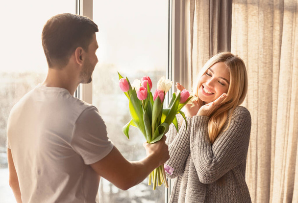 Romantic young man presenting flowers to his girlfriend at home - Фото, изображение