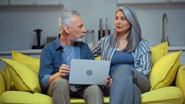 ηλικιωμένο διαφυλετικό ζευγάρι μιλάει κοντά σε φορητό υπολογιστή στο σαλόνι  - Πλάνα, βίντεο