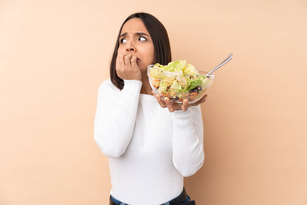 Young brunette girl holding a salad over isolated background nervous and scared putting hands to mouth - Photo, Image