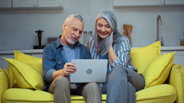 ηλικιωμένο διαφυλετικό ζευγάρι μιλάει κοντά σε φορητό υπολογιστή στο σαλόνι  - Πλάνα, βίντεο
