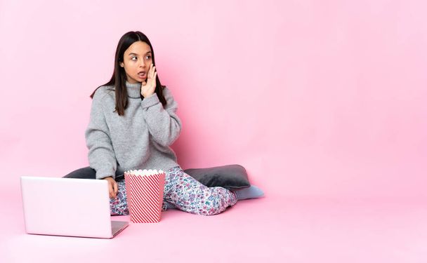 Young mixed race woman eating popcorn while watching a movie on the laptop whispering something with surprise gesture while looking to the side - Fotografie, Obrázek