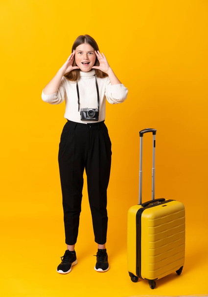 Full body of traveler teenager girl with suitcase over isolated yellow background shouting with mouth wide open - Fotografie, Obrázek