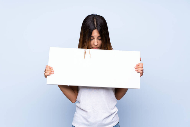 Jonge vrouw over geïsoleerde blauwe achtergrond met een leeg wit bordje voor invoegen van een concept - Foto, afbeelding