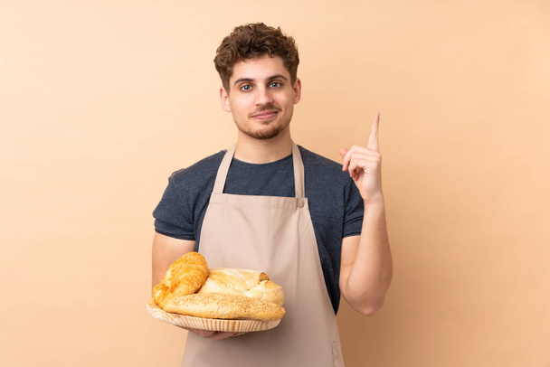 Panadero masculino sosteniendo una mesa con varios panes aislados sobre fondo beige apuntando con el dedo índice una gran idea
 - Foto, imagen