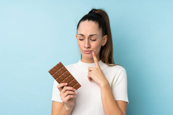 jonge brunette vrouw over geïsoleerde blauwe achtergrond het nemen van een chocolade tablet en met twijfels - Foto, afbeelding