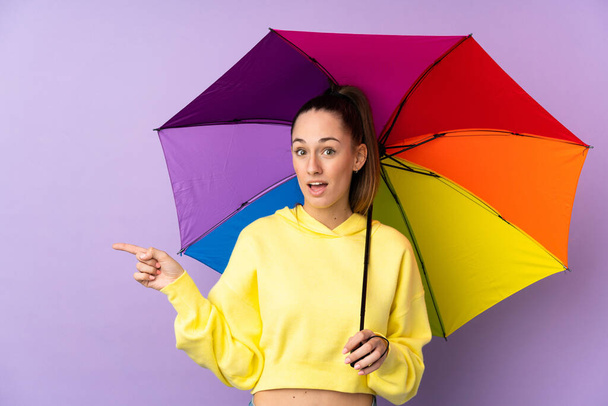 Mladá brunetka žena drží deštník nad izolovanou fialovou stěnou překvapený a ukazuje prstem na stranu - Fotografie, Obrázek