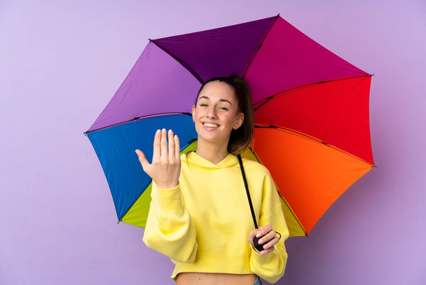 Młoda brunetka trzymająca parasol nad odizolowaną, purpurową ścianą, zapraszająca do pomocy. Cieszę się, że przyszedłeś. - Zdjęcie, obraz