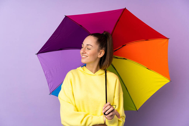 Mladá brunetka žena drží deštník nad izolovanou fialovou stěnou se šťastným výrazem - Fotografie, Obrázek