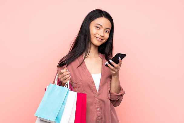 Teenager ragazza cinese isolata su sfondo rosa tenendo borse della spesa e scrivendo un messaggio con il suo cellulare ad un amico - Foto, immagini