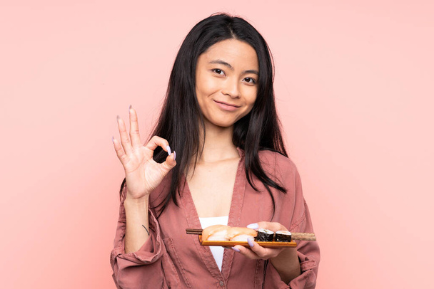 Έφηβη Ασιάτισσα που τρώει σούσι απομονωμένη σε ροζ φόντο που δείχνει μια πινακίδα εντάξει με δάχτυλα - Φωτογραφία, εικόνα