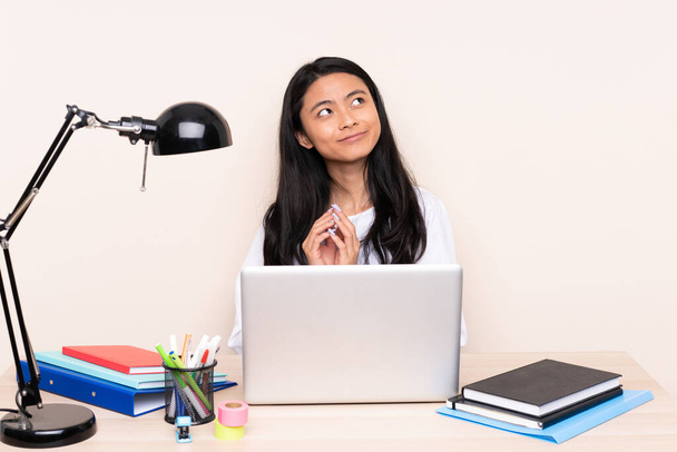 Студентка азиатка на рабочем месте с ноутбуком, изолированным на бежевом фоне, замышляющая что-то
 - Фото, изображение