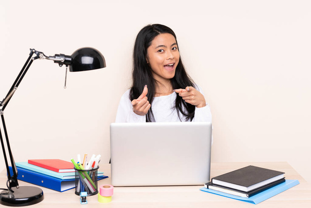 Estudante menina asiática em um local de trabalho com um laptop isolado no fundo bege apontando para a frente e sorrindo
 - Foto, Imagem