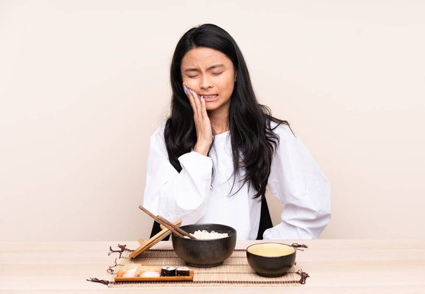Teenager Asian girl eating asian food isolated on beige background with toothache - Фото, изображение