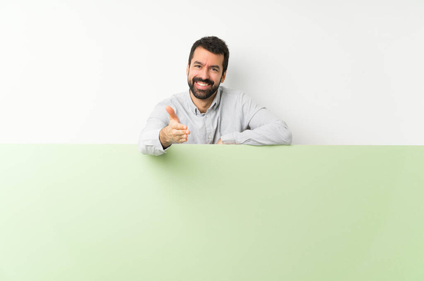 Jovem homem bonito com barba segurando um grande cartaz verde vazio apertando as mãos para fechar um bom negócio
 - Foto, Imagem