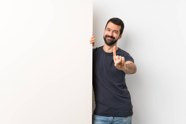 Joven hombre guapo con barba sosteniendo un gran cartel vacío mostrando y levantando un dedo
 - Foto, Imagen