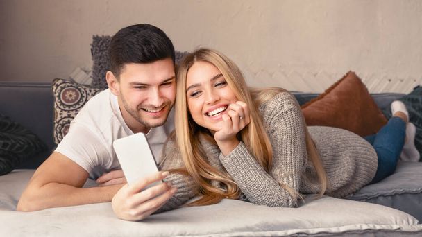 Loving young couple with mobile phone lying on sofa at home - Fotografie, Obrázek