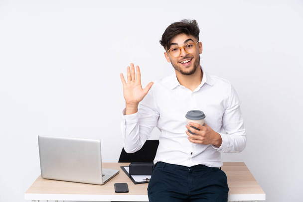 Hombre árabe en una oficina aislado sobre fondo blanco saludando con la mano con expresión feliz
 - Foto, Imagen