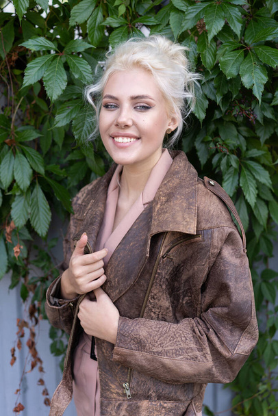 Счастливая блондинка в кожаной куртке, улыбающаяся рядом с растениями
 - Фото, изображение