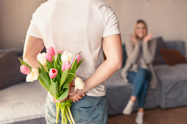 Young man hiding bouquet of tulips for girlfriend behind his back at home, blank space - Fotografie, Obrázek
