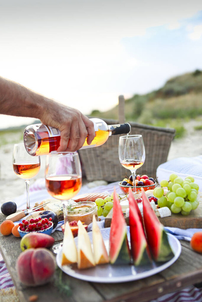 Crop male serving pink wine having picnic with delicious snacks and fruit on sandy beach - Fotó, kép