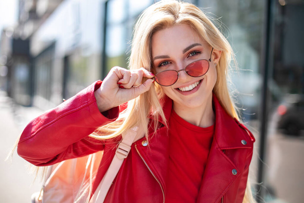 Closeup selfie-portrait student of attractive girl in sunglasses with long hairstyle and snow-white smile in city. happy blonde wearing red leather jacket - Foto, Imagen