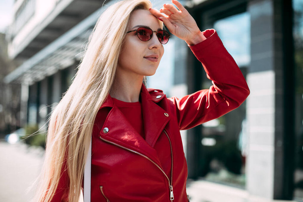 Closeup selfie-portrait student of attractive girl in sunglasses with long hairstyle in city.happy blonde wearing red leather jacket. - Zdjęcie, obraz