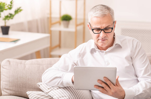 Homme âgé utilisant une tablette numérique assis sur le canapé
 - Photo, image