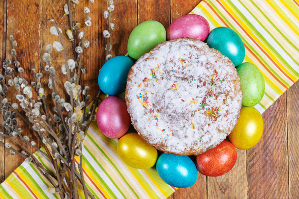 Easter decorations, background. Homemade Easter cake with colored eggs around it and willow branches on striped kitchen towel on wooden table. Top view. - Photo, Image