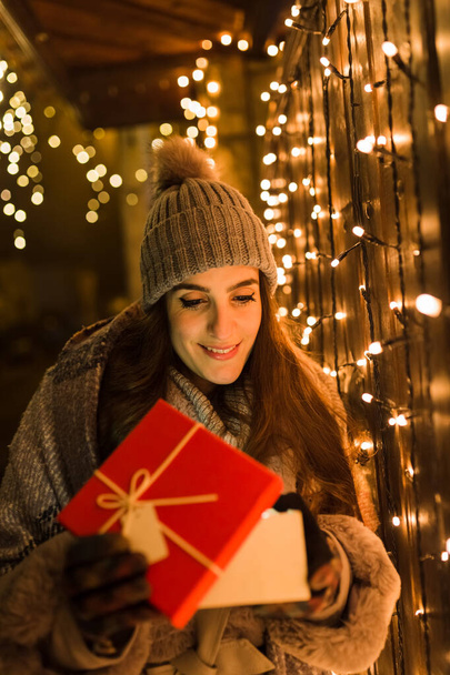 Smiling woman opening christmas present gift box next to fence decorated with holiday lights on the street. - Fotografie, Obrázek