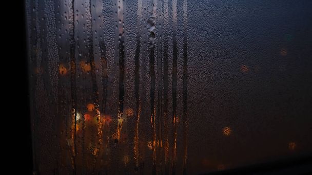 Detailní záběr okna s kapkami deště na rozmazaném pozadí světel. Koncept. Tmavá světla prosvítají tmavým oknem rozmazaným kapkami deště - Fotografie, Obrázek