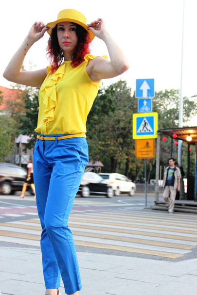 woman with red curly hair, in blue classic trousers, a yellow blouse and a yellow hat stands on a pedestrian crossing in city environments - Fotoğraf, Görsel