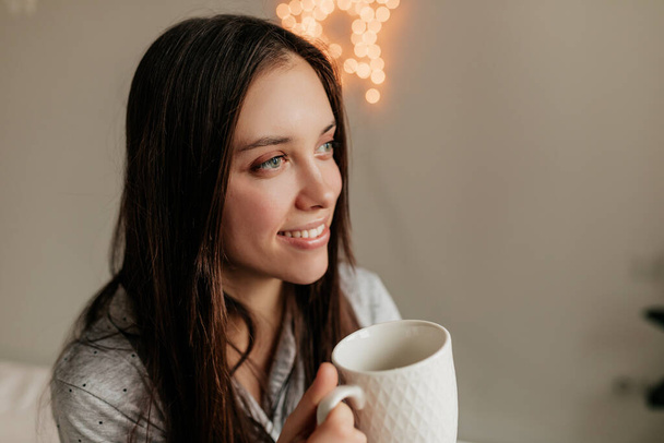 Κοντινό πλάνο εσωτερική φωτογραφία του χαμογελαστού ευτυχισμένη γυναίκα με σκούρα μαλλιά κοιτάζοντας το παράθυρο, ενώ πίνοντας καφέ - Φωτογραφία, εικόνα