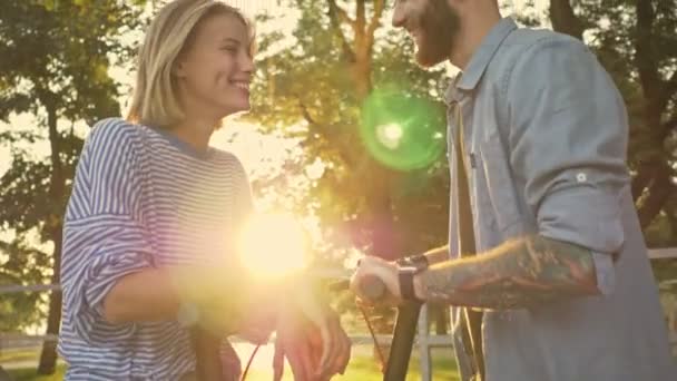 Wesoła młoda para śmieje się i rozmawia podczas jazdy skuterami elektrycznymi w zielonym parku o zachodzie słońca - Materiał filmowy, wideo