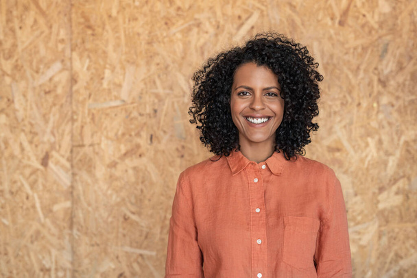 Portrait of a smiling young woman with curly hair standing against a chipboard wall in her workshop - Foto, Bild