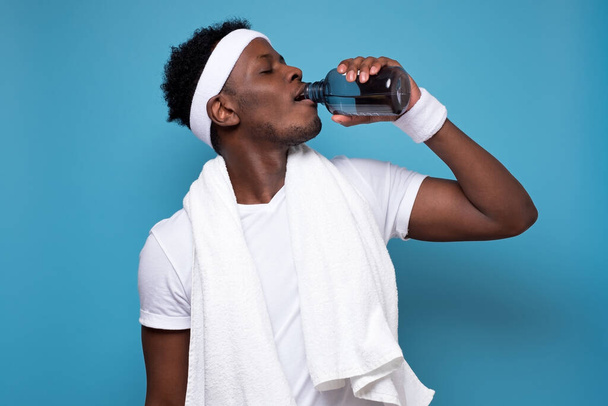Young african sportsman drinking water after training. Studio shot on blue wall. - Photo, Image