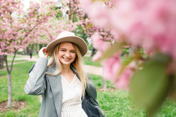 young beautiful blonde girl in a white dress, a gray cloak and a pink hat stands in a park near sakura trees - Foto, immagini