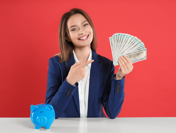 Young woman with money and piggy bank at table on crimson background - Foto, Imagen